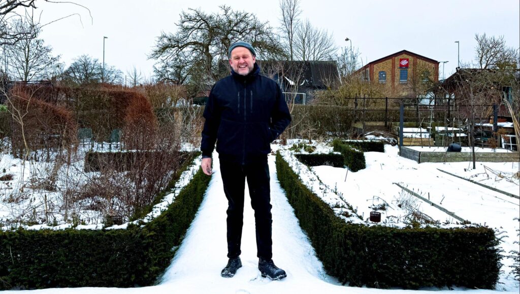 Leende medelålders man i svart jacka och mössa står på en snöig gång i en trädgård.