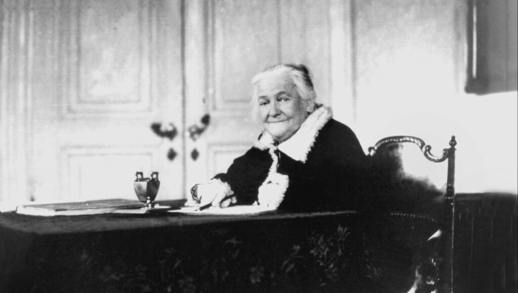 Svartvitt porträtt av den äldre kvinnan Clara Zetkin sittande vid ett skrivbord i en sal.