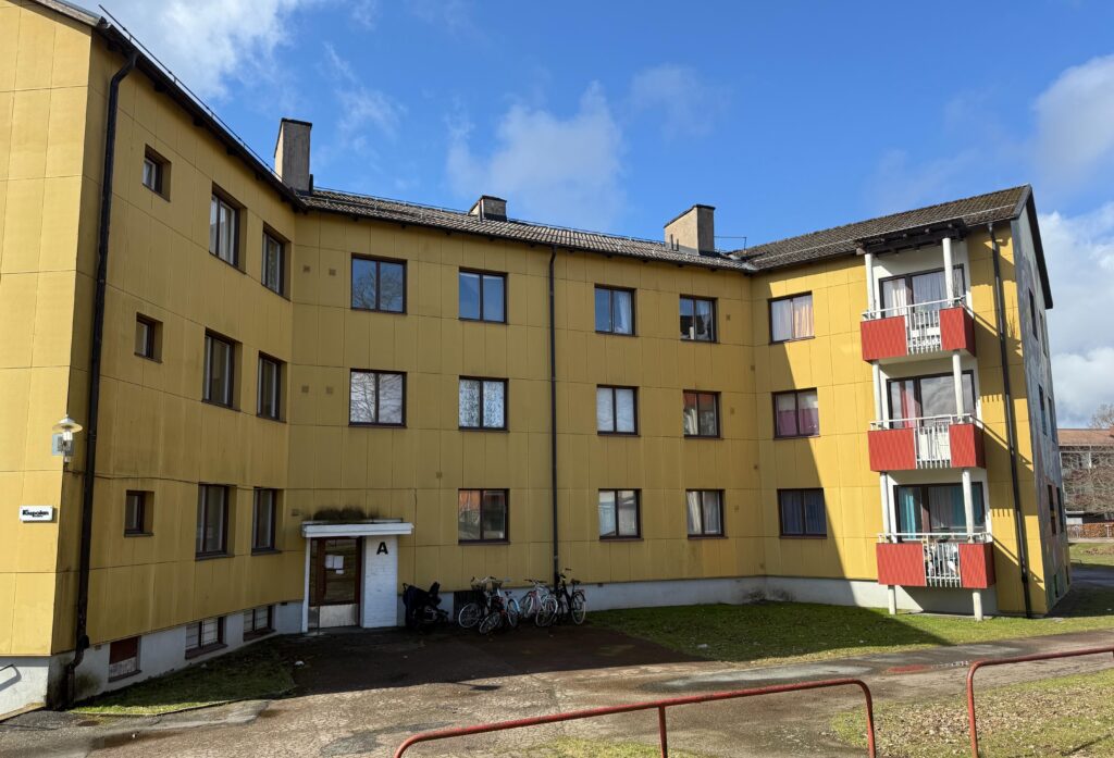 Tre våningar högt gult hyreshus med röda balkonger och parkerade cyklar vid entrén under en klarblå himmel.