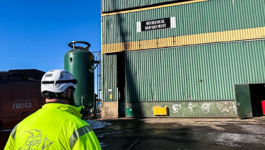 Arbetare i neongul varseljacka och vit hjälm står framför en stor grön plåtbyggnad på ett industriområde.