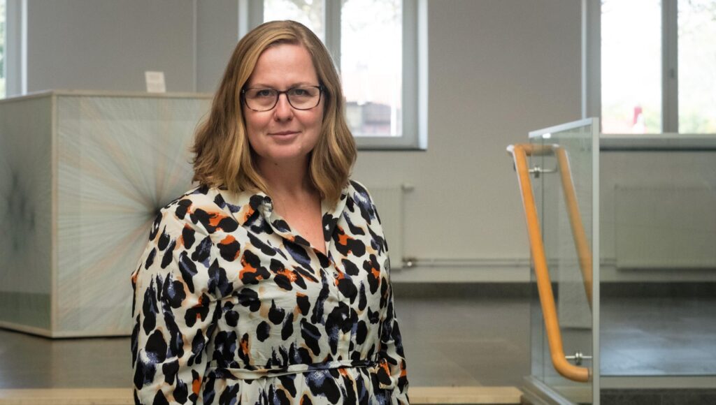 Kvinna i glasögon och leopardmönstrad blus står i en ljus inomhusmiljö med trappa i bakgrunden