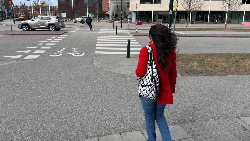 Kvinna i röd kavaj och mönstrad ryggsäck sedd bakifrån vid ett övergångsställe i stadsmiljö.
