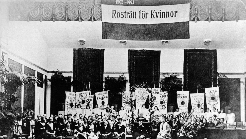 Svartvit bild på en stor grupp kvinnor framför fanor och en banderoll där det står Rösträtt för Kvinnor.