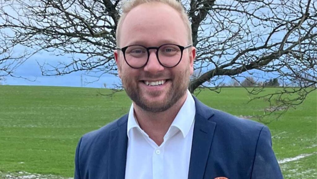 Skeende man med glasögon och blå kavaj framför en grön åker och ett träd utan löv.