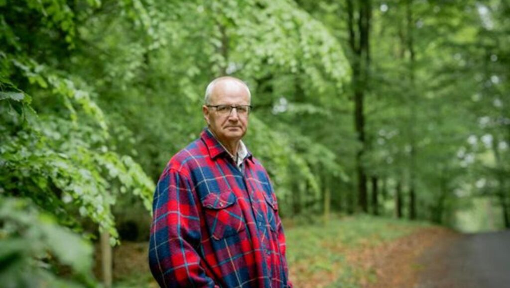 En man med glasögon och rutig flanellskjorta står i en grönskande lövskog.