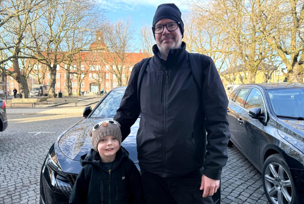 En man i svart jacka och ett barn i stickad mössa står framför en mörk bil på en öppen plats.