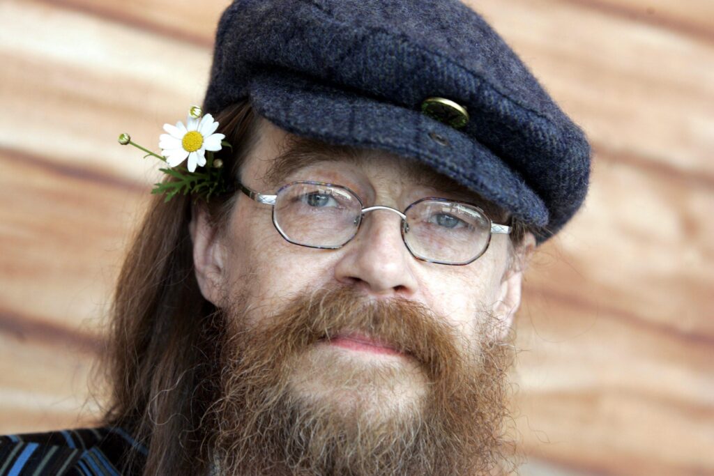 Närbild på Peps Persson med skägg, keps, glasögon och en blomma bakom örat.