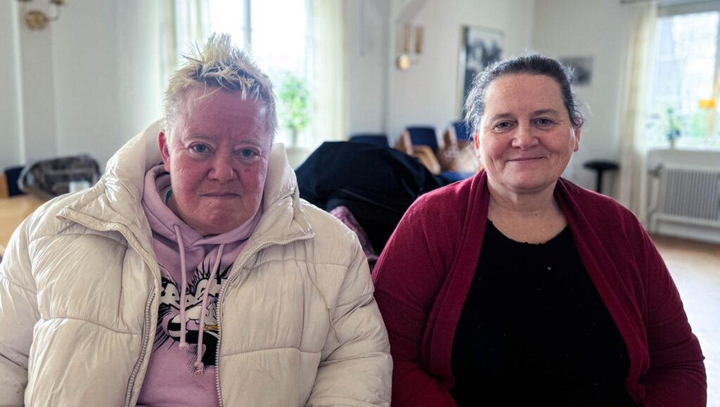 Två kvinnor sitter bredvid varandra inomhus, en i vit täckjacka och en i röd kofta.