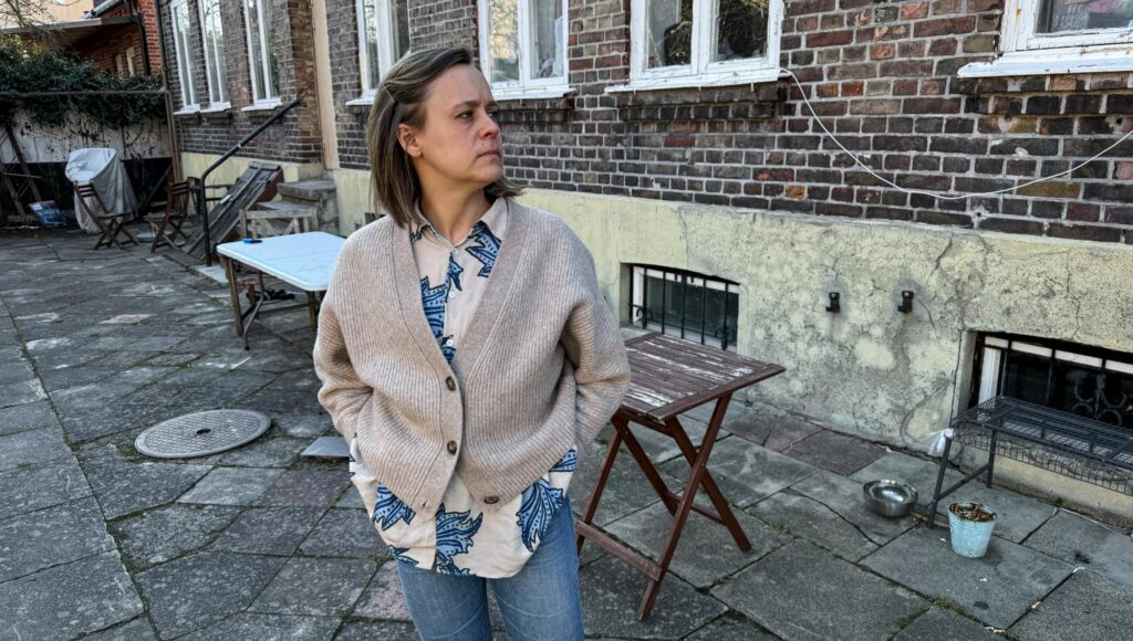 Kvinna i stickad kofta står på en stenlagd innergård framför en tegelvägg