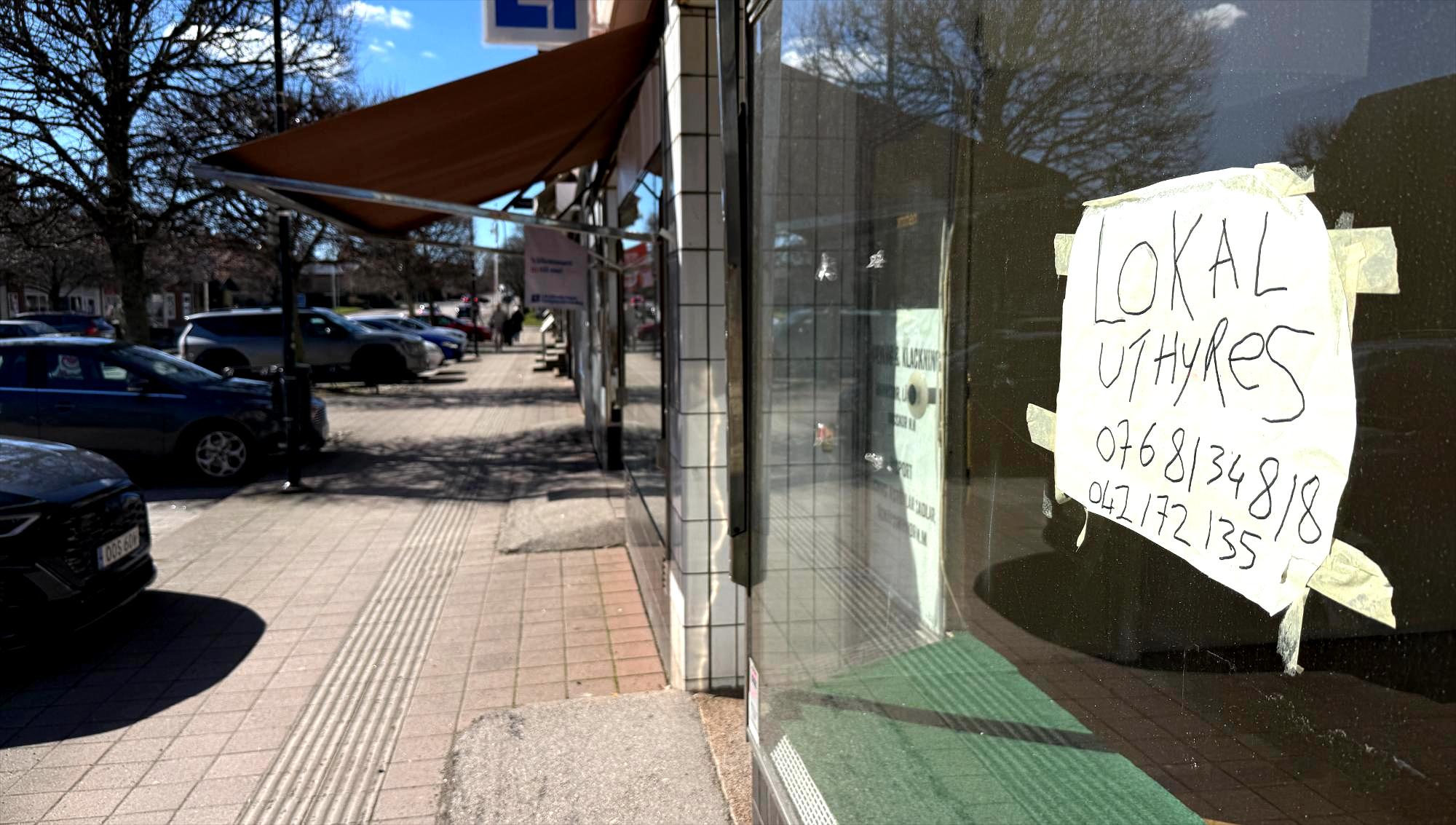 En handskriven lapp med texten 'Lokal uthyres' sitter upptejpad på ett skyltfönster längs en solig butiksgata i Åstorp.