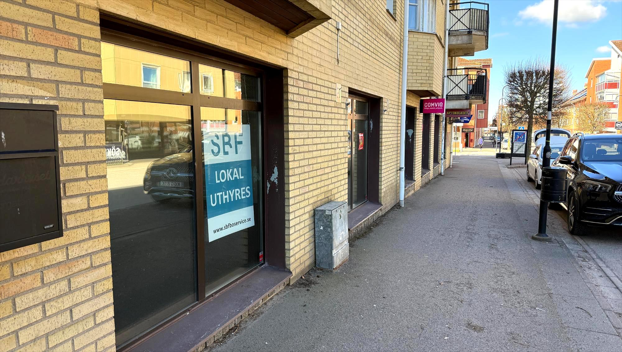 En tom affärslokal med en skylt om uthyres sitter i fönstret på en gul tegelbyggnad längs en trottoar i Åstorp.