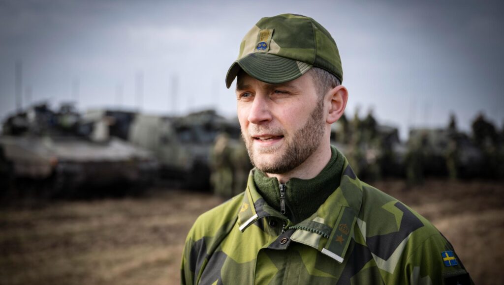 Man i svensk militäruniform och keps utomhus med pansarfordon i suddig bakgrund.