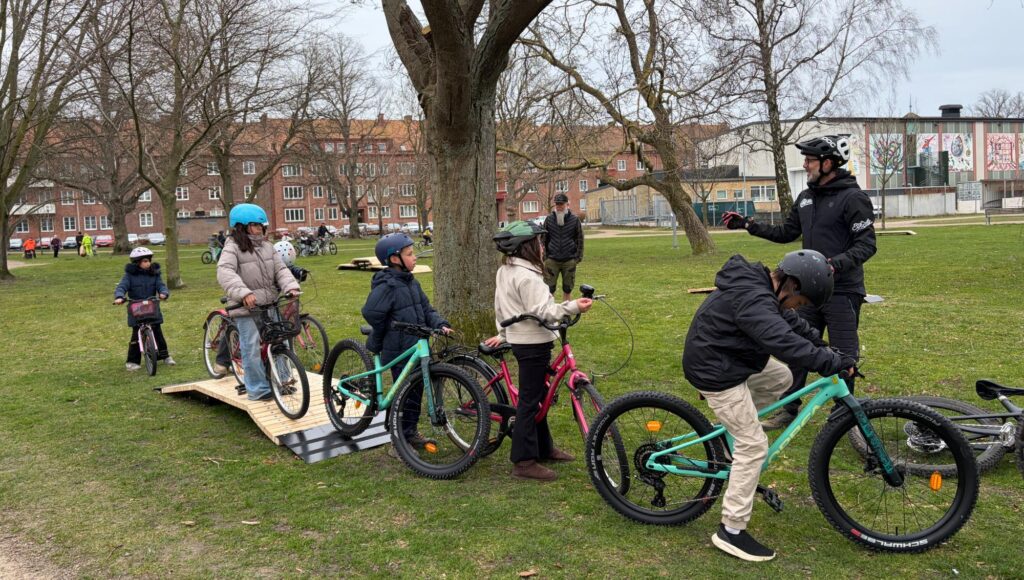 Barn med cyklar och hjälmar övar på träramper i en park tillsammans med en vuxen instruktör.
