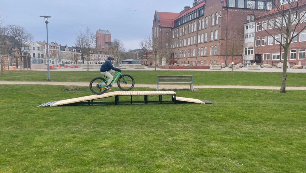 En ung person i hjälm cyklar på en träramp i en park framför en stor tegelbyggnad.