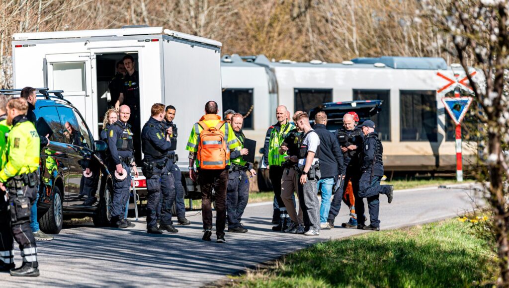 Polispersonal och räddningsarbetare samlade kring fordon och en tågvagn vid en järnvägsövergång.