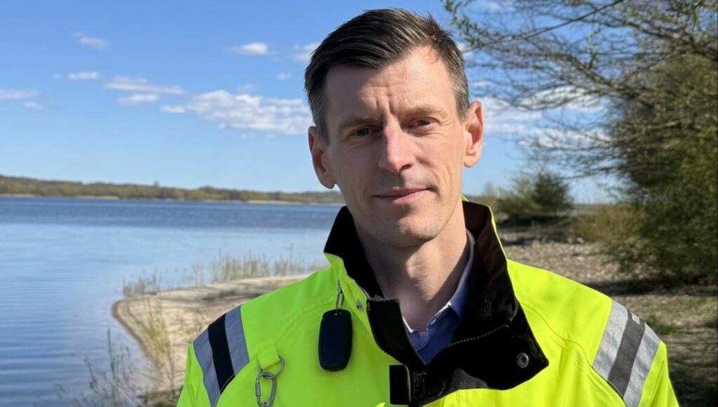 En man i 40-årsåldern i neongul varseljacka står vid en strandkant under en blå himmel.