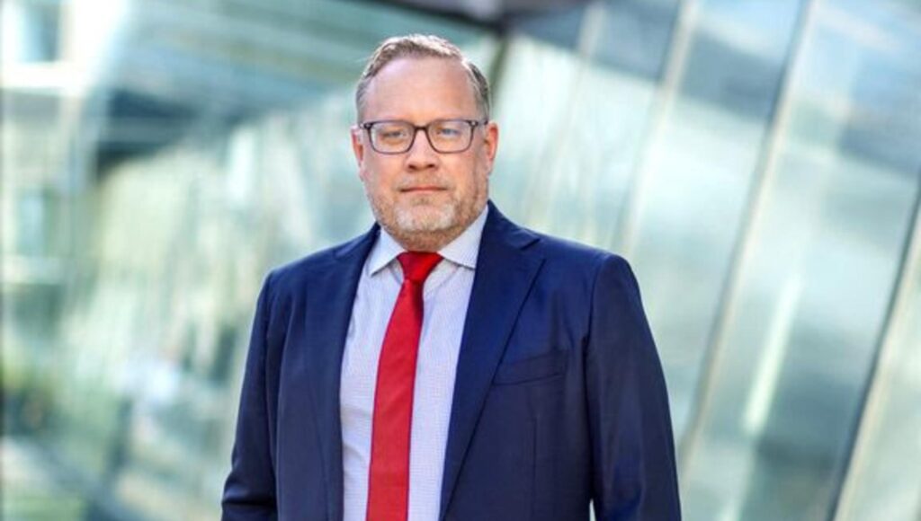 Man i 50-årsåldern med glasögon, blå kavaj och röd slips mot en ljus glasfasad i bakgrunden.