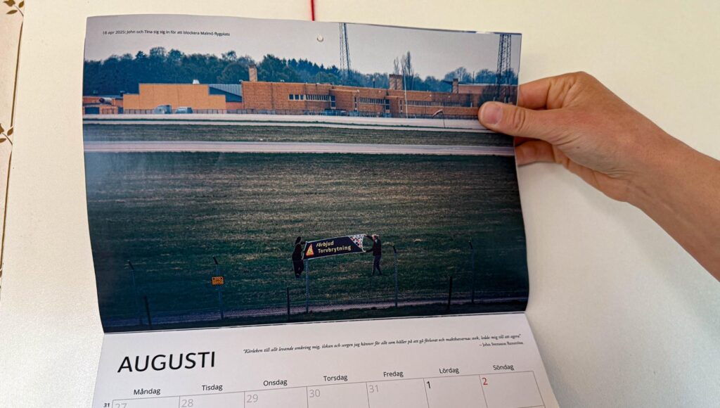 En person håller upp en kalender som visar två personer med en banderoll på ett flygfält framför en gul byggnad.