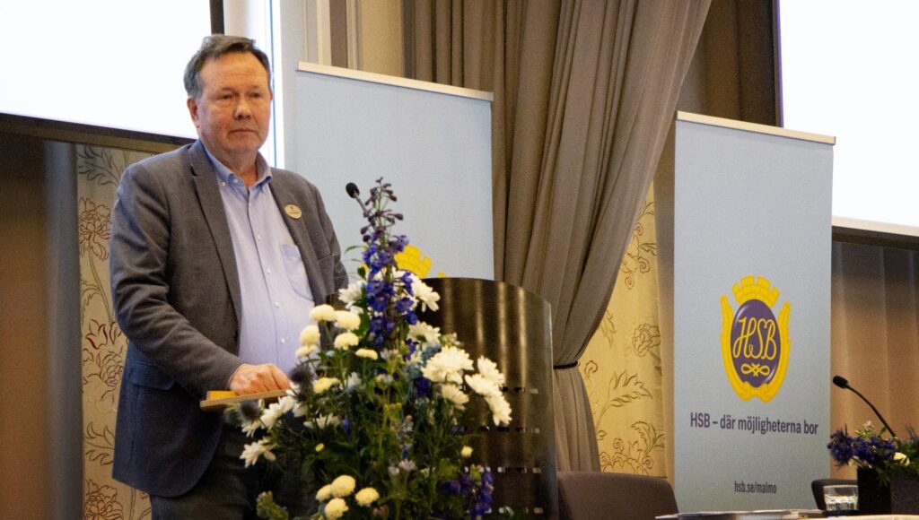 En man i 60-årsåldern står bakom en talarstol med blommor framför HSB-banderoller.