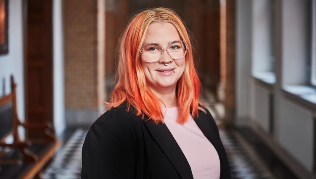 Kvinna i 30-årsåldern med rödorange hår, glasögon och svart kavaj ler mot kameran.