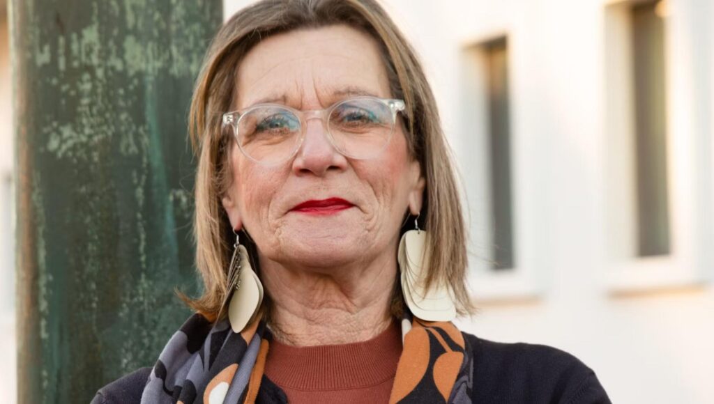 Porträtt på kvinna med glasögon och stora örhängen vid en grönmålad pelare.