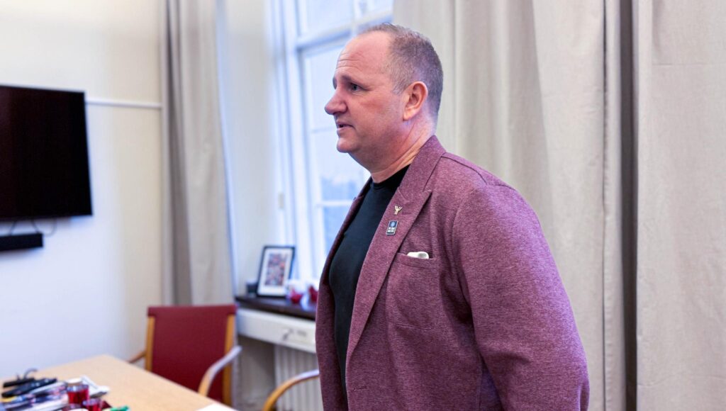 Man i lila kavaj står i profil i ett ljust rum med fönster och gardiner i bakgrunden.