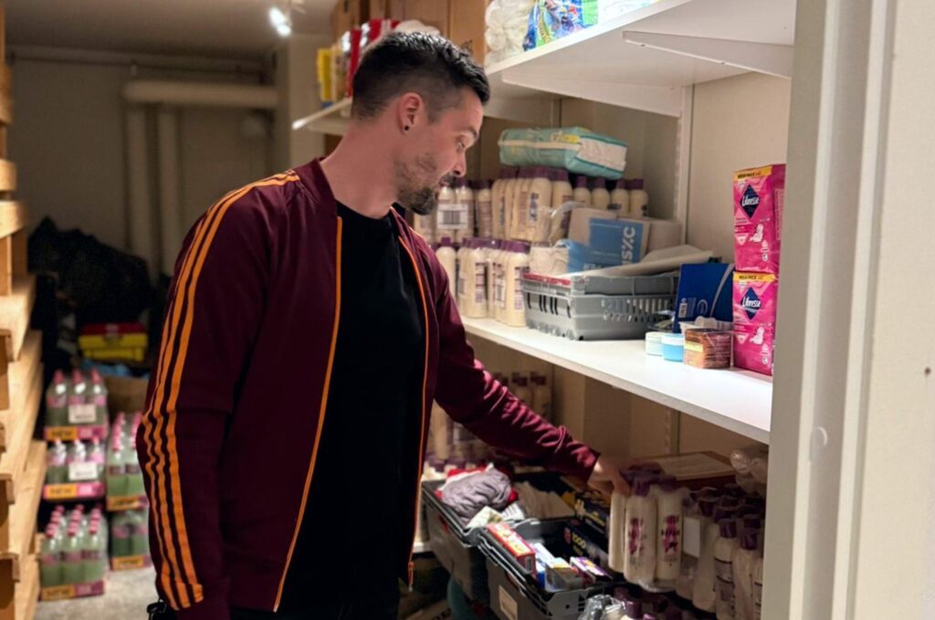 En man i vinröd träningsjacka sorterar flaskor på en hylla i ett välfyllt förråd.