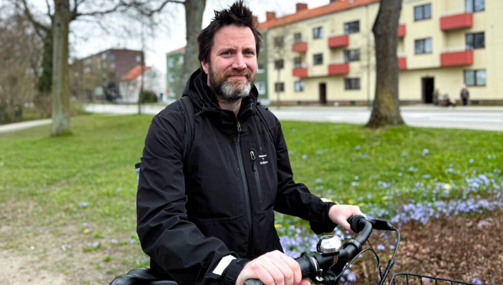 Man med skägg och svart jacka håller i ett cykelstyre utomhus framför flerbostadshus.