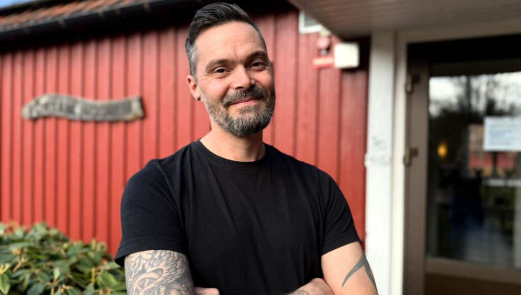 Leende man i 40-årsåldern med skägg och tatueringar framför en röd trävägg.