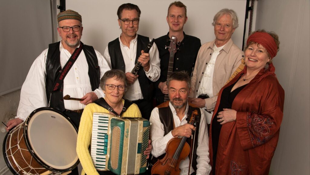 Sju personer i olika åldrar ler och håller i musikinstrument mot en vit fond.