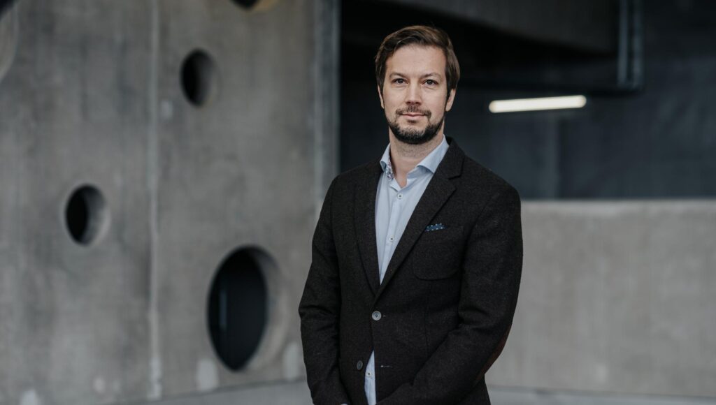 En man i 40-årsåldern med skägg bär mörk kavaj framför en grå betongvägg med runda hål.