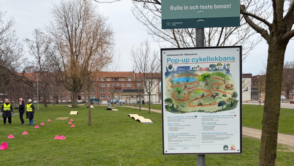 Informationsskylt om en pop-up cykellekbana på en gräsyta med träningshinder i bakgrunden.