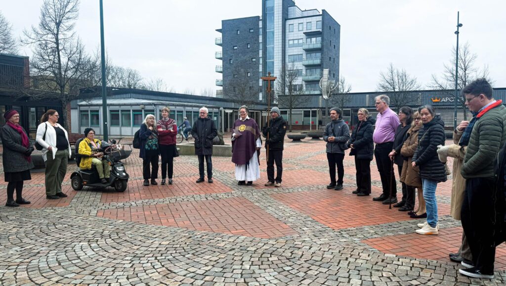En grupp människor står i en halvcirkel på ett torg kring en präst och ett kors.