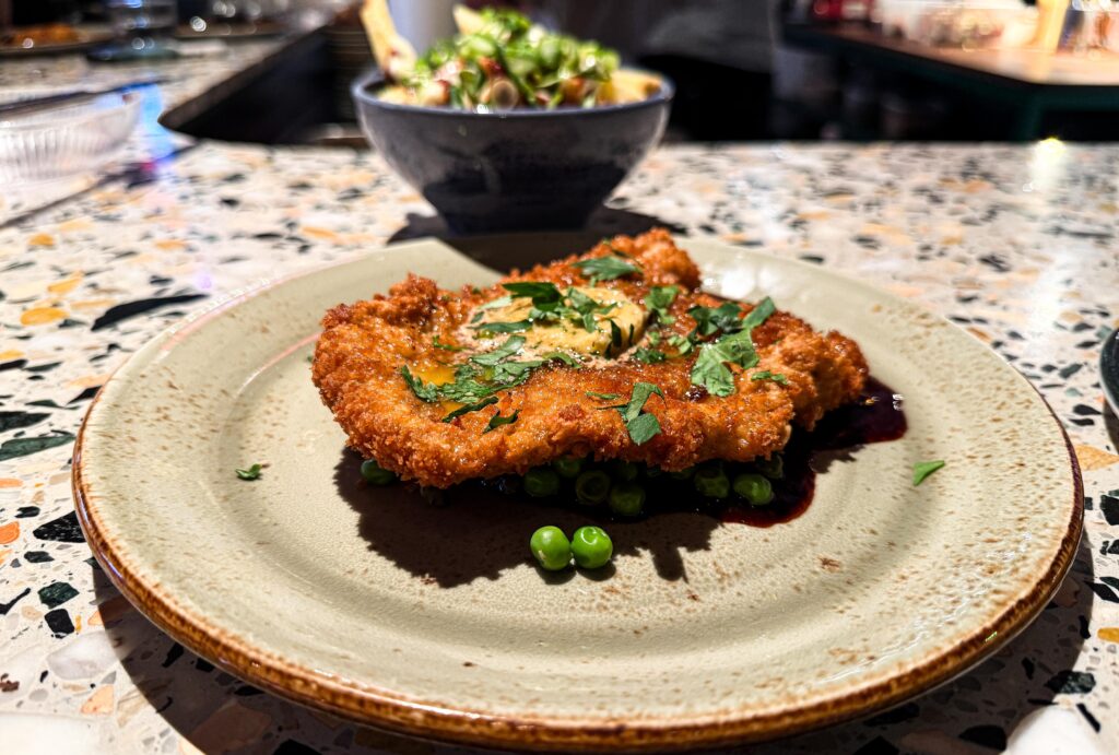 Panerad schnitzel med örtsmör och gröna ärtor på en tallrik i en restaurangmiljö.