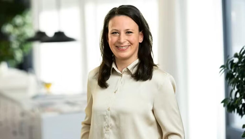 Leende kvinna med mörkt hår i ljus skjorta fotograferad i en ljus kontorsmiljö med växter.
