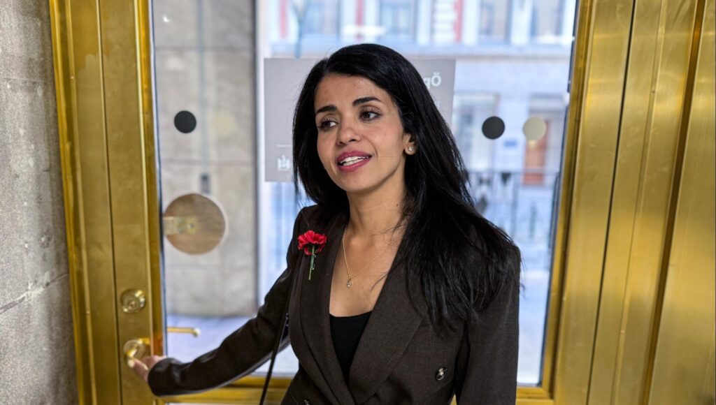 Anfal Mahdi i mörk kavaj med en röd nejlika på slaget står vid en guldfärgad dörr.