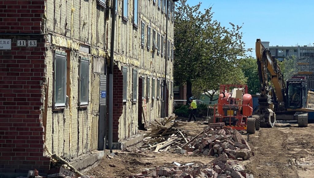 Ett bostadshus med frilagd isolering intill en byggarbetsplats med tegelrester och grävmaskiner.