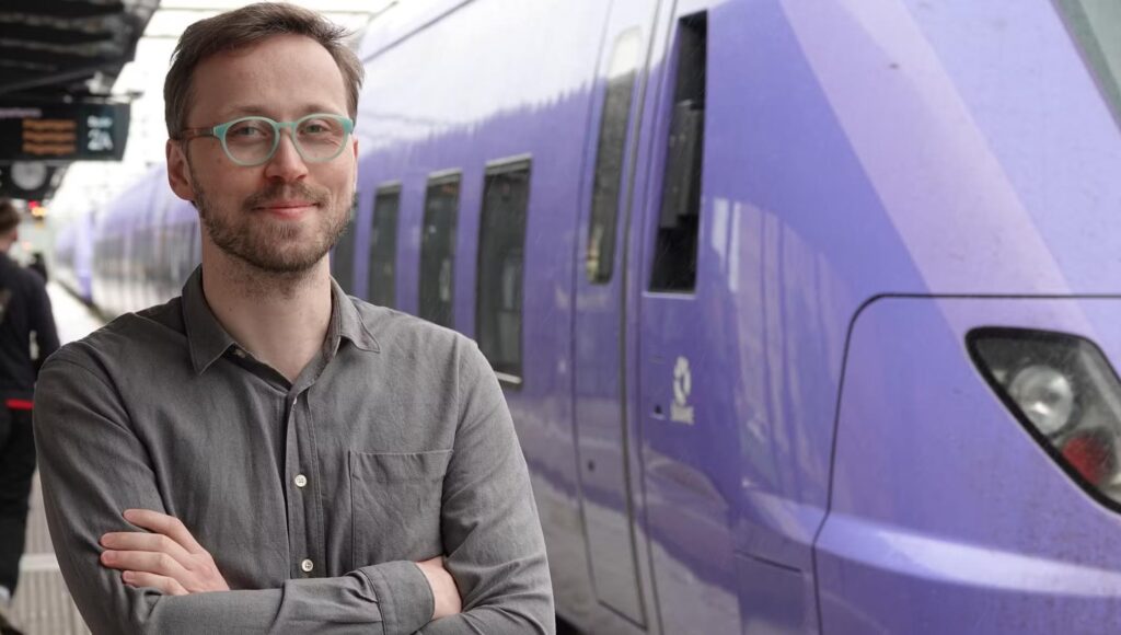 Man med glasögon står med armarna i kors framför ett lila tåg på en perrong