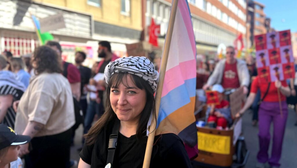 Kvinna i keffiyeh håller en transflagga under ett demonstrationståg i stadsmiljö.