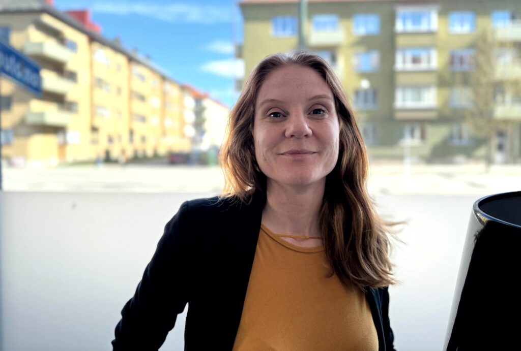 Långhårig kvinna i 35-årsåldern ler mot kameran framför ett suddigt bostadsområde i dagsljus.