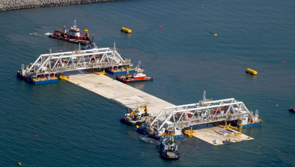 Flygbild på stora vita fackverkskonstruktioner som flyttar ett betongelement i havet med hjälp av bogserbåtar.