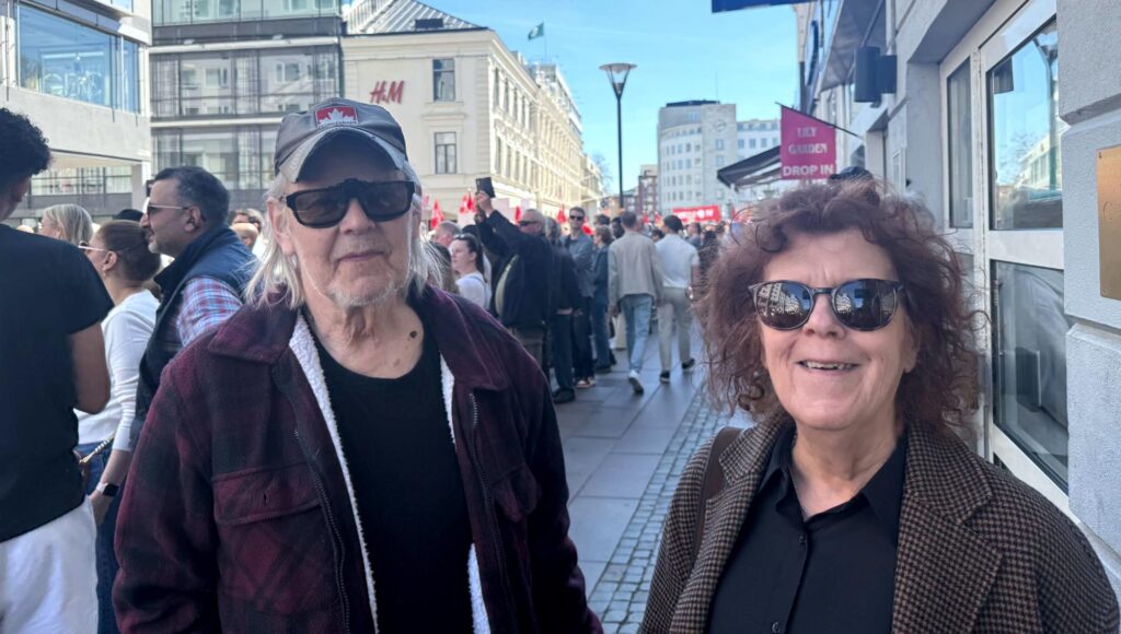 En äldre man i keps och en kvinna med rufsigt hår ler mot kameran på en folktät stadsgata i solljus.