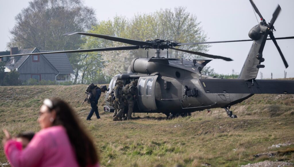 Svart militärhelikopter på ett gräsfält där uniformerad personal med ryggsäckar kliver ombord nära ett bostadshus.