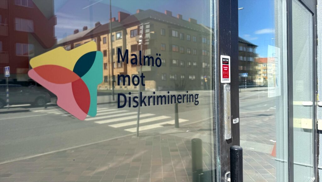 En glasdörr med logotypen och texten Malmö mot Diskriminering där stadsmiljön speglas i glaset.