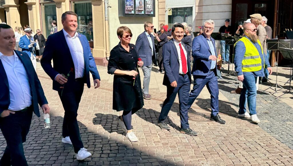 Personer i formell klädsel med röda rosor promenerar på en kullerstensgata i solsken.