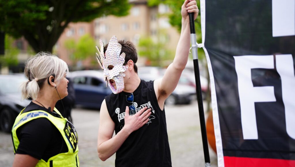Maskerad demonstrant med flagga samtalar med polis i varselväst.