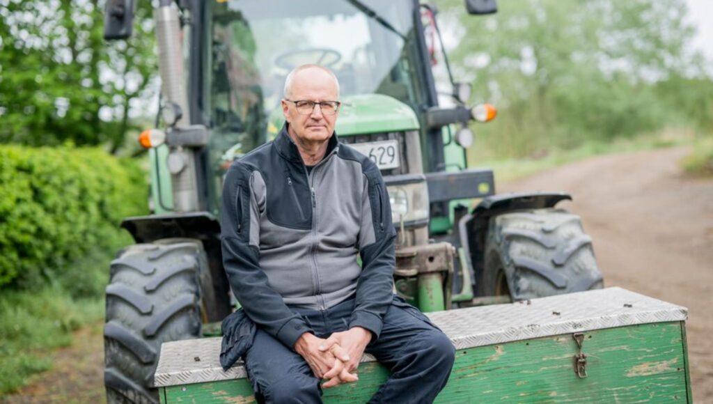 En man i 60-årsåldern sitter på en grön trälåda framför en stor traktor på en grusväg.