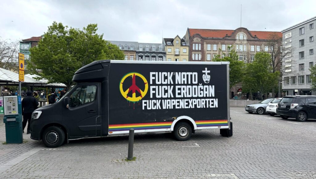 Svart lätt lastbil med politisk text mot Nato, Erdogan och vapenexport parkerad på ett kullerstenstorg.