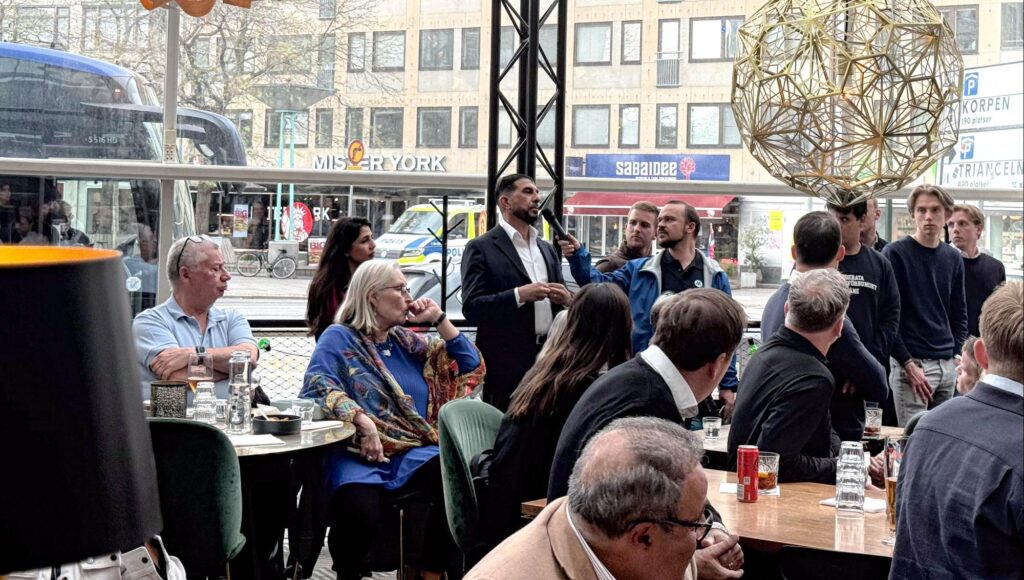 En man talar i mikrofon inför en sittande publik i en lokal med stora fönster mot en stadsgata.