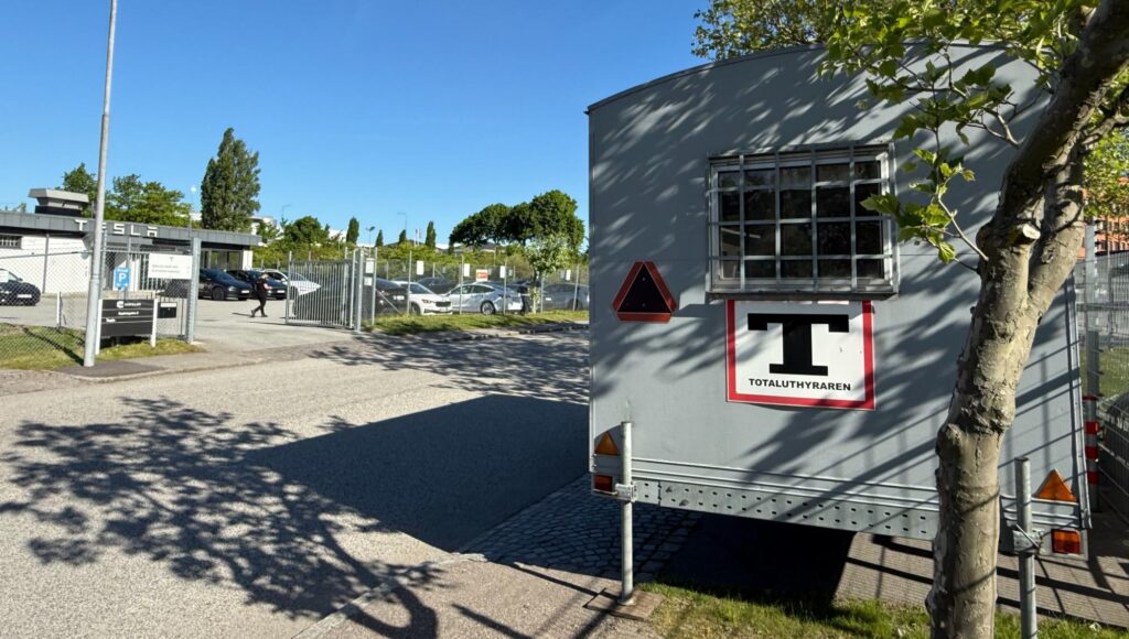 En grå arbetsvagn med logotypen Totaluthyraren står parkerad intill en Tesla-anläggning i solsken.
